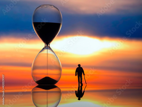 miniature people. silhouette of an elderly man walking towards the sunset next to an hourglass. end of life. photo