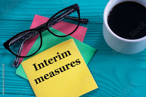 Billede på lærred INTERIM MEASURES - words on yellow note sheets on green background with a cup of