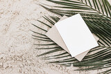 Summer stationery still life. Closeup of blank card mock-up and craft envelope on green date palm leaves. Sandy beach background or desert. Flat lay, top view. Tropical vacation concept.