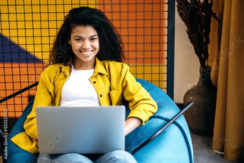 Bilde på lerret Happy curly girl student or freelancer is sitting in chair at modern office, using laptop