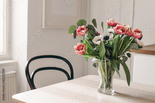 Spring flowers in glass vase on wooden table. Blurred kitchen background with old chair. Bouquet of red tulips, white anemone flowers and eucalyptus branches. Contemporary elegant, Scandinavian