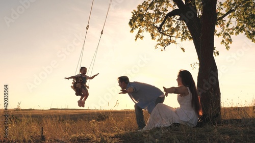 Parents swing the little girl on a swing on a summer day, flying like an airplane in the sky, happy mother and father daughter play together outdoors, family travel on vacation or vacation, teamwork