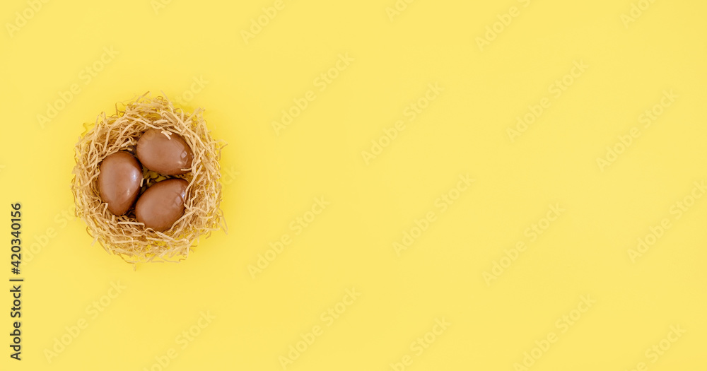 A flat lay or top view of chocolate eggs in nest on the yellow background. Easter hunting. Copy space, place for text, banner. Color of the year illuminating.