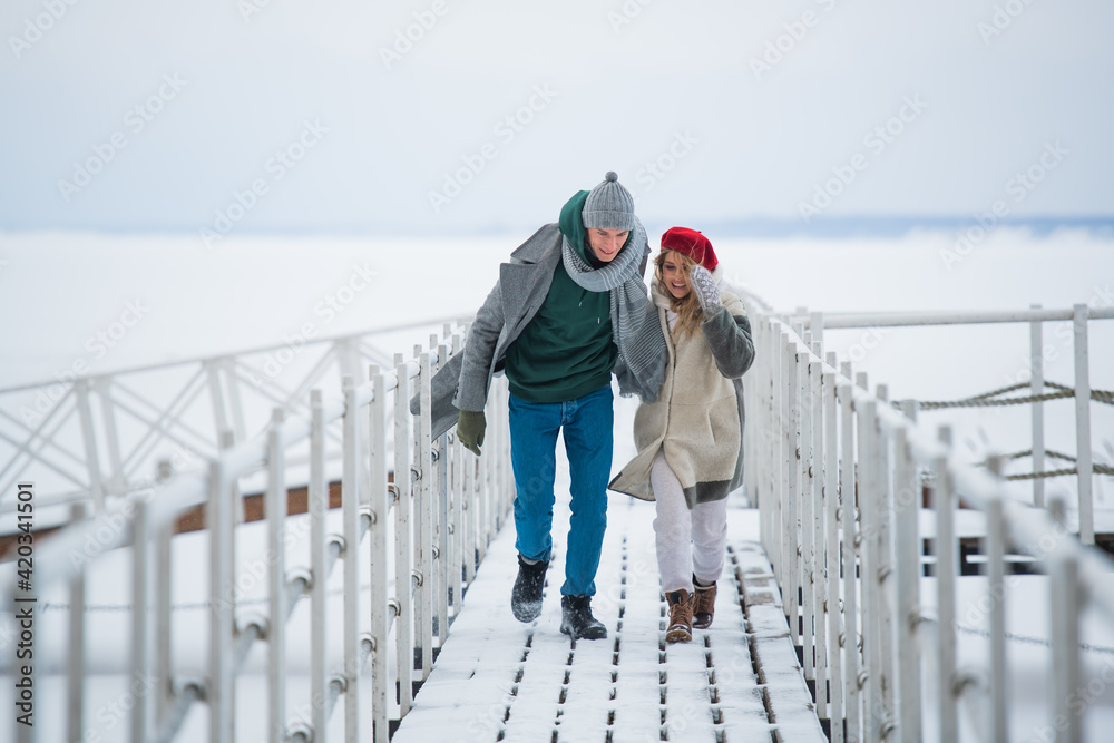 A young man and woman spend time together on a winter vacation, walking on a snow-covered pier on the river.