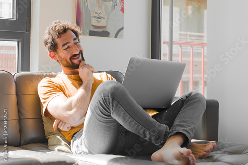 young bearded man smiling with a happy, confident expression with hand on chin, wondering and looking to the side with a laptop on a couch laptop concept