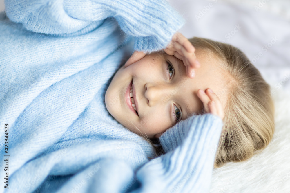 Cute little caucasian girl seven years old with blonde hair lying on bed at home in bedroom. Kid wearing stylish cozy knitted sweater blue color. Happy Child smiling Cute little caucasian girl seven years old with blonde hair lying on bed at home in bedroom. Kid wearing stylish cozy knitted sweater blue color. Happy Child smiling