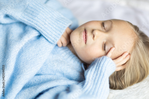Cute little caucasian girl seven years old with blonde hair lying on bed at home in bedroom. Kid wearing stylish cozy knitted sweater blue color. Happy Child smiling