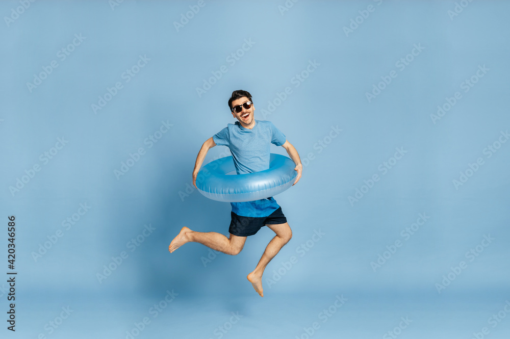 Full length photo of a joyful happy caucasian guy in a sunglasses, a blue t-shirt and with a blue inflatable ring, jumping high on a isolated blue background, travel, beach vacation concept