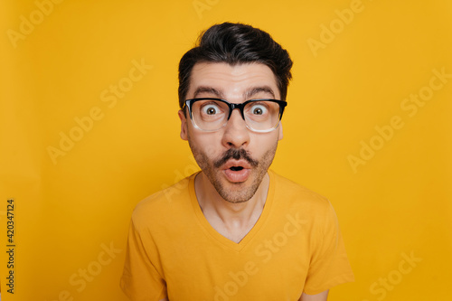 Portrait of amazed foolish cheerful caucasian guy in glasses and in an orange t-shirt, cheerfully looking at the camera with a distorted face, standing against an isolated orange background
