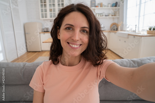 Happy vlogger woman recording new content to vlog. Dark-eyed millennial girl with curly hair wear t-shirt smiling broadly, making selfie, stretching arm to camera, photographing herself at home. 