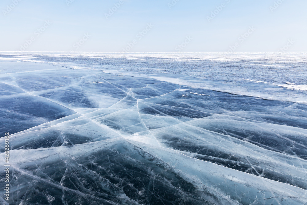 Fototapeta premium Beautiful icy background of frozen lake Baikal surface on a cold winter day. Blue transparent ice with cracks. Cold icy endless desert. Unusual winter landscape. Natural blue pattern