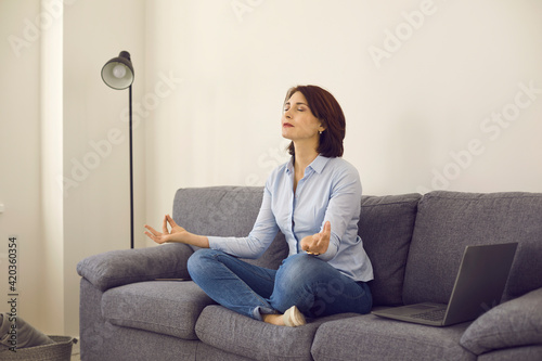 Caucasian middle age self-employed woman entrepreneur meditating doing breathing exercise sitting in lotus yoga position on home sofa. Mental health recovery, mindfulness and conditioning concept