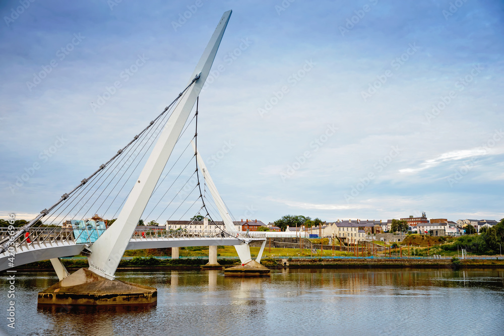 Fototapeta premium Derry, Ireland. Illuminated Peace bridge in Derry Londonderry, City of Culture, in Northern Ireland with city center at the background. Night cloudy sky with reflection in the river at the dusk