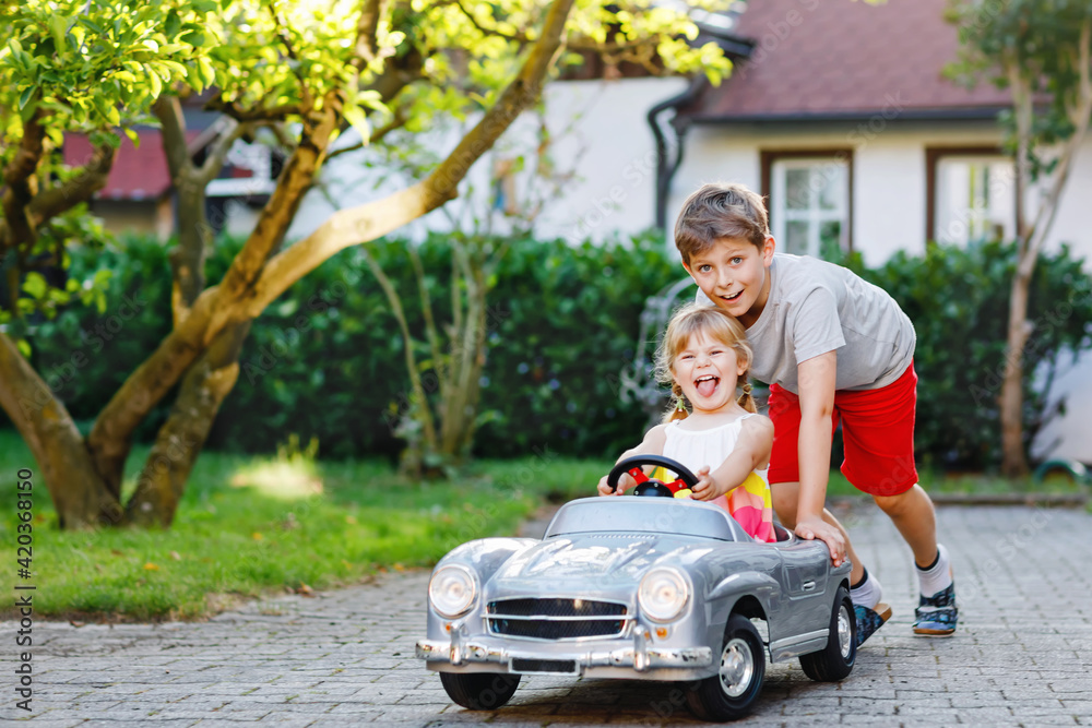 Two happy children playing with big old toy car in summer garden, outdoors. Kid boy pushing and driving car with little toddler girl, cute sister inside. Laughing and smiling kids. Lovely family Two happy children playing with big old toy car in summer garden, outdoors. Kid boy pushing and driving car with little toddler girl, cute sister inside. Laughing and smiling kids. Lovely family