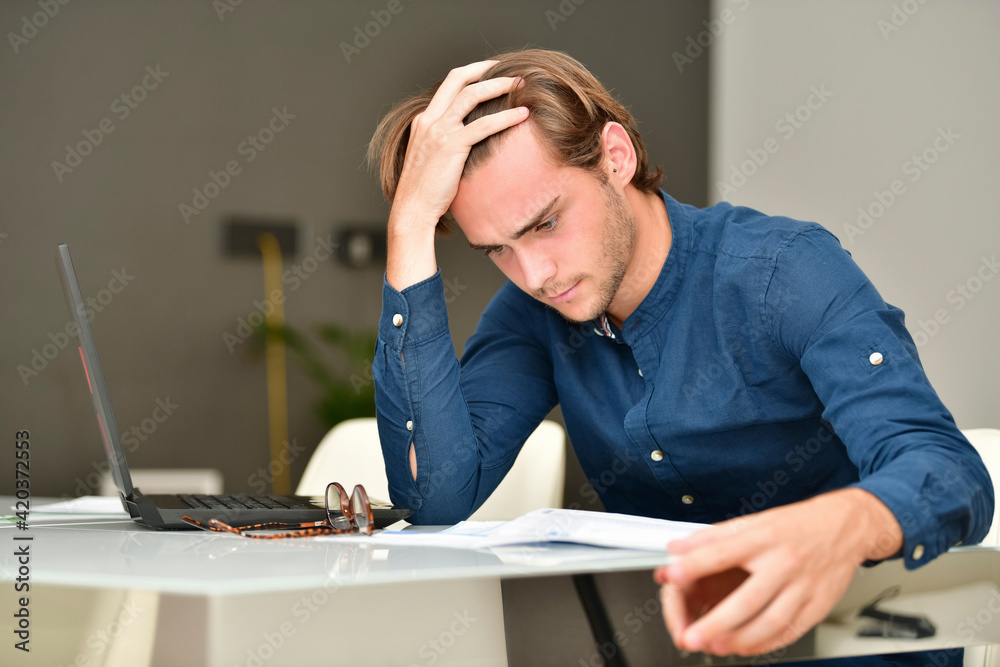 handsome young man looking closely to some bills on a table next to a laptop. Payments and technology concept. handsome young man looking closely to some bills on a table next to a laptop. Payments and technology concept.