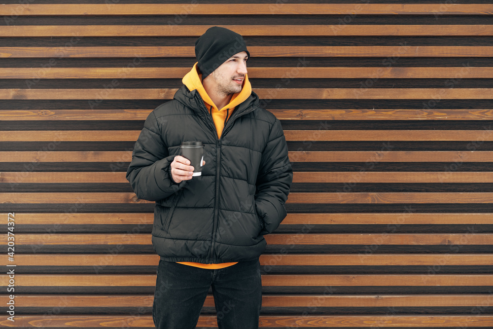 Young man in winter clothes standing with coffee to go in a street against wooden wall Young man in winter clothes standing with coffee to go in a street against wooden wall