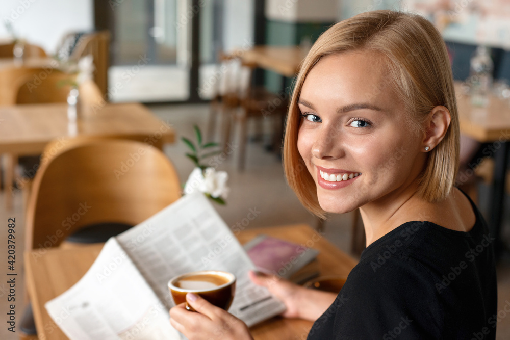 Modern beautiful blond woman reading newspaper while drinking coffee at coffee shop interior, internet distance work