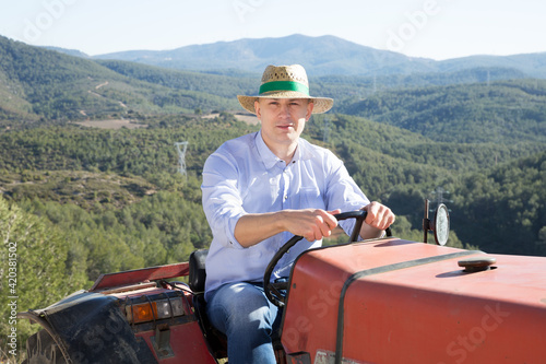 Portrait of adult male in straw hat sitting on tractor on background with vineyard