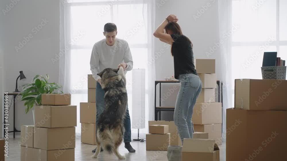 Long shot. The young family is happy to move to a new home. A young man is dancing with his dog. A cheerful woman in a T-shirt and jeans is dancing next to them.