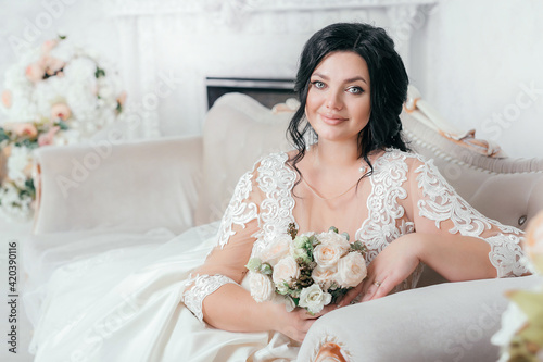 Beautiful sexy brunette bride plus size in a light photo studio. Wedding photo. Plus size model with beautiful makeup and hairstyle. Curvy girl bride with a veil. Wedding bouquet