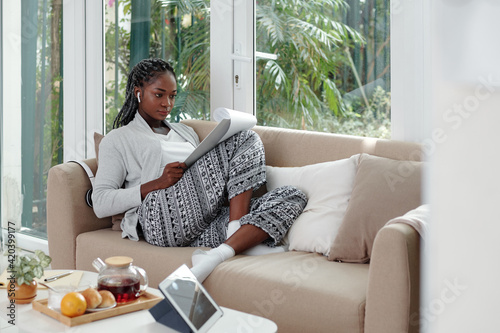 Concentrated Black young woman in loungewear working from home, she is sitting on sofa and reading business document photo