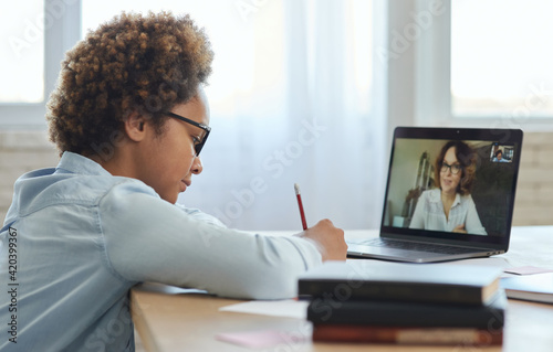 Focused mixed race teen schoolgirl listening to her teacher, making notes during online video lesson while studying from home