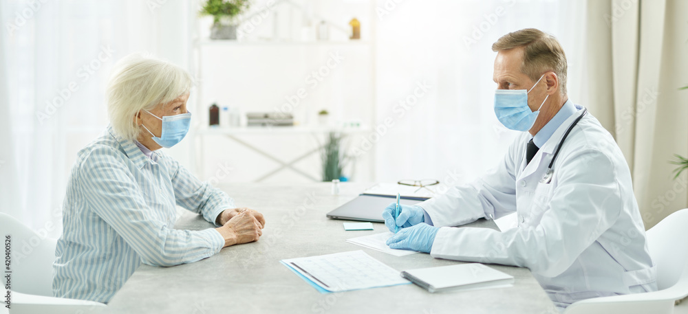Fototapeta premium Doctor in medical mask sitting at the table and talking with old lady