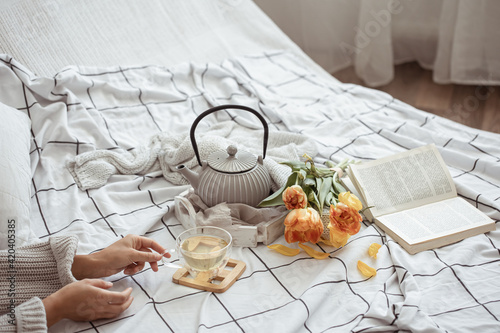 Spring home composition in bed with tea in female hands, a teapot, a book and a bouquet of flowers.