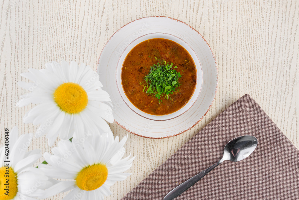 Tomato soup with herbs on a white plate on a light wooden table against a background of chamomile flowers. Tomato soup with herbs on a white plate on a light wooden table against a background of chamomile flowers.