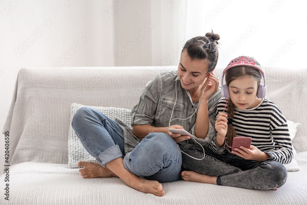 A young mother and her daughter are listening to music on their phones while sitting on the couch at home.