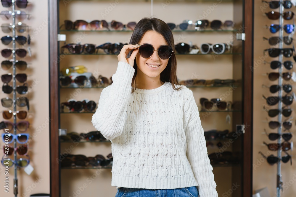 Which one is better fit me. Portrait of cheerful hesitating woman in optician store, making decision, holding stylish sunglasses, choosing what she should buy. Great discounts for buying two pairs
