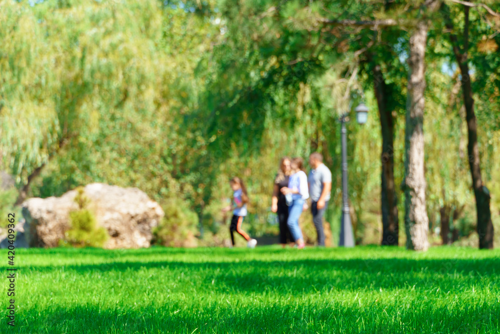 Obraz premium people walking and children playing in a city park on a summer day, green lawns with grass and trees, paths and benches, bright sunlight and shadows