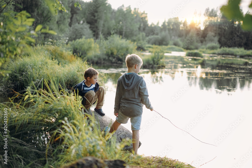 two caucasian boys pretending fishing on the pond. One is holding a cane imegine it is a rod. Image with slective focus and backlight