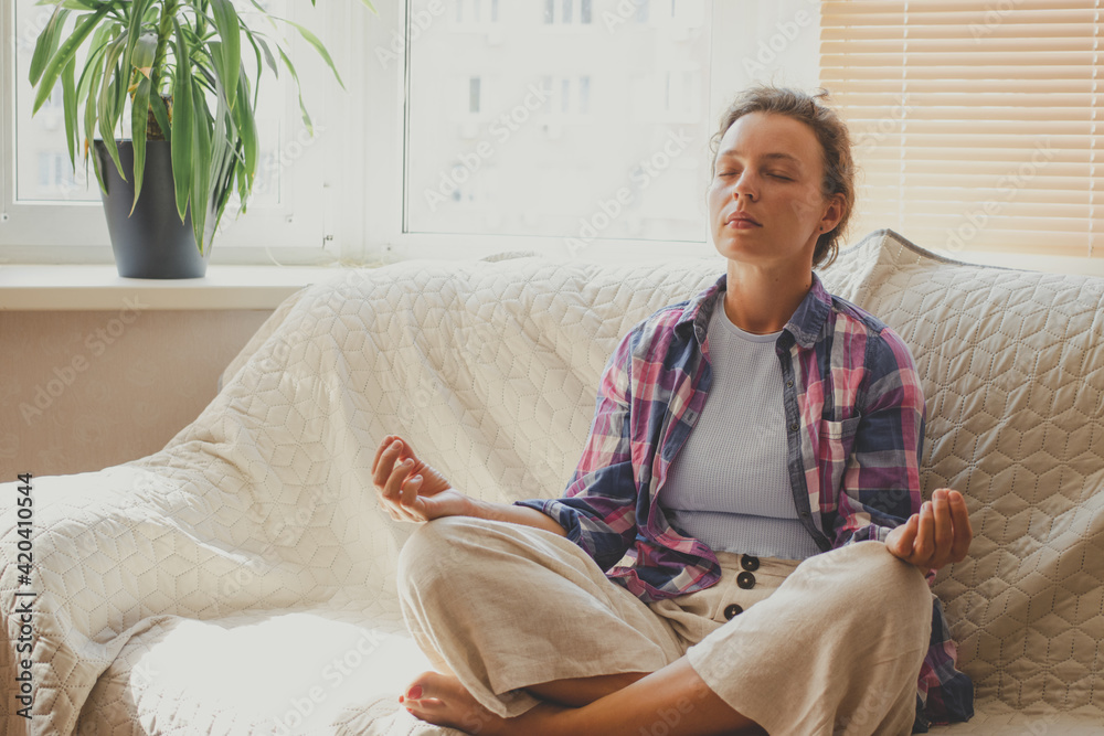 Fototapeta premium Young woman sitting on couch at home meditating with closed eyes, wearing casual cloth. Doing yoga exercises, breathing practices to calm down mind, relieve tension and stress. Lotus pose. Copy space