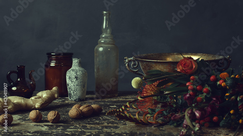 Old worn wooden table with two glass bottles, a glass pitcher, a glass jar, a bronze bowl, ginger and walnuts and flowers.
