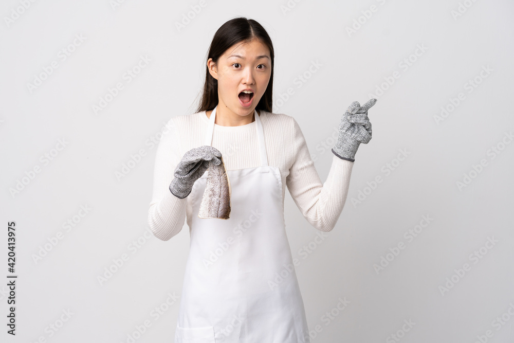 Chinese fishmonger wearing an apron and holding a raw fish over isolated white background surprised and pointing finger to the side Chinese fishmonger wearing an apron and holding a raw fish over isolated white background surprised and pointing finger to the side