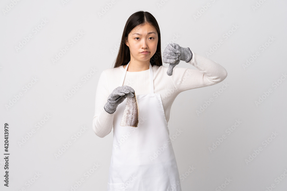 Chinese fishmonger wearing an apron and holding a raw fish over isolated white background showing thumb down with negative expression Chinese fishmonger wearing an apron and holding a raw fish over isolated white background showing thumb down with negative expression