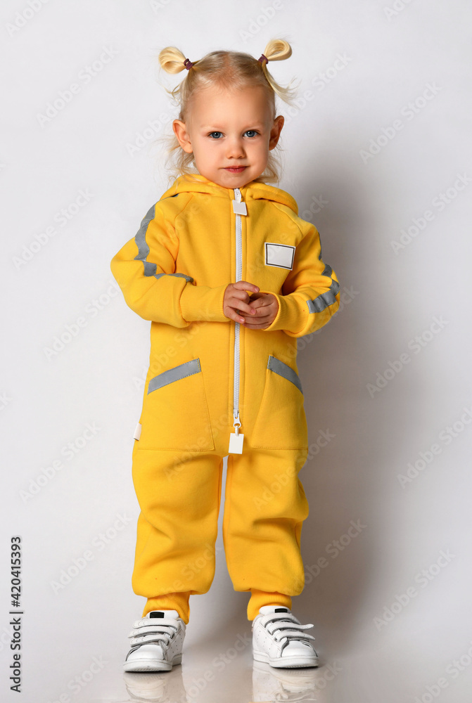 Little cute blonde girl in a warm comfortable overalls standing against a gray wall. Happy childhood and fashionable baby clothes Little cute blonde girl in a warm comfortable overalls standing against a gray wall. Happy childhood and fashionable baby clothes