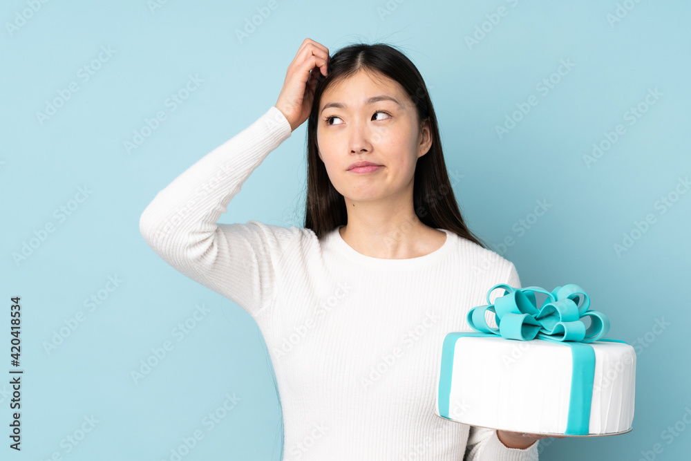Pastry asian chef holding a big cake isolated on blue background having doubts and with confuse face expression