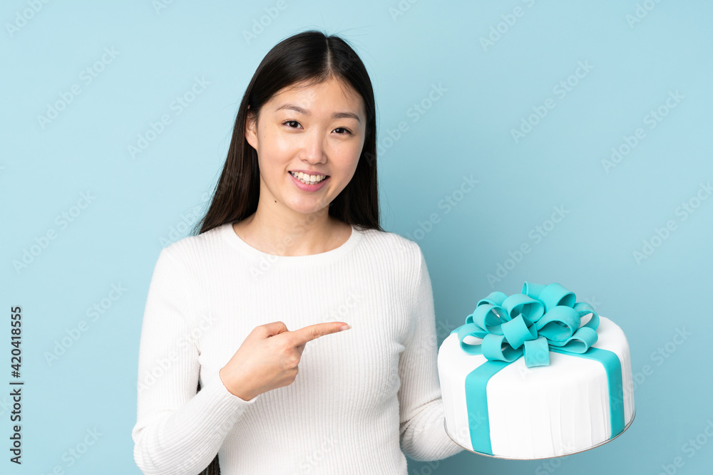 Pastry asian chef holding a big cake isolated on blue background and pointing it Pastry asian chef holding a big cake isolated on blue background and pointing it
