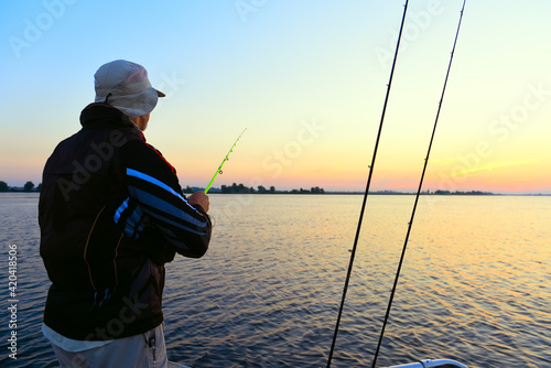 A male fisherman with a spinning rod in his hand catches fish from a boat at dawn.