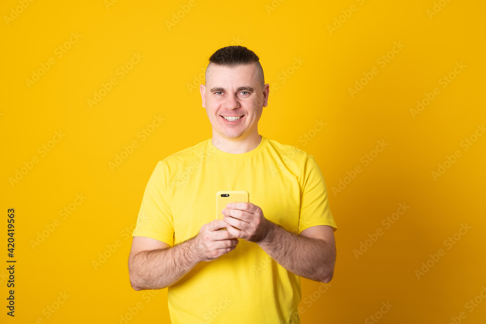 Young man texting message on smartphone isolated on yellow background. Smiling man holding smartphone and looking at it. Happy man writing a message on the phone. Young man texting message on smartphone isolated on yellow background. Smiling man holding smartphone and looking at it. Happy man writing a message on the phone.