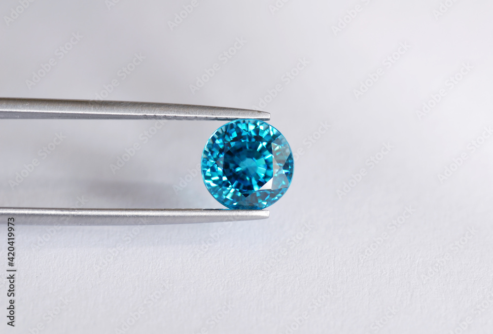 Natural rich electric blue round bullet faceted in Thailand heated zircon clean gemstone in tweezers on daylight from window. Light gray gradient background. Natural rich electric blue round bullet faceted in Thailand heated zircon clean gemstone in tweezers on daylight from window. Light gray gradient background.