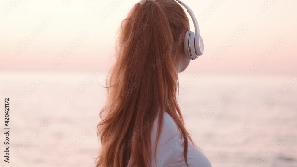 A pleased young redhead woman wearing headphones is looking around while walking outside near the sea in the morning A pleased young redhead woman wearing headphones is looking around while walking outside near the sea in the morning