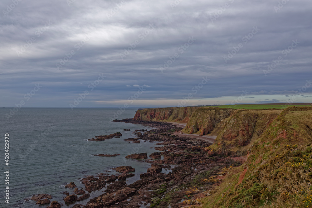 Shingle Beaches, rocky outcrops and Red Sandstone Cliffs with Farmland above seen from the Angus Coastal Footpath under a dark sky. Shingle Beaches, rocky outcrops and Red Sandstone Cliffs with Farmland above seen from the Angus Coastal Footpath under a dark sky.