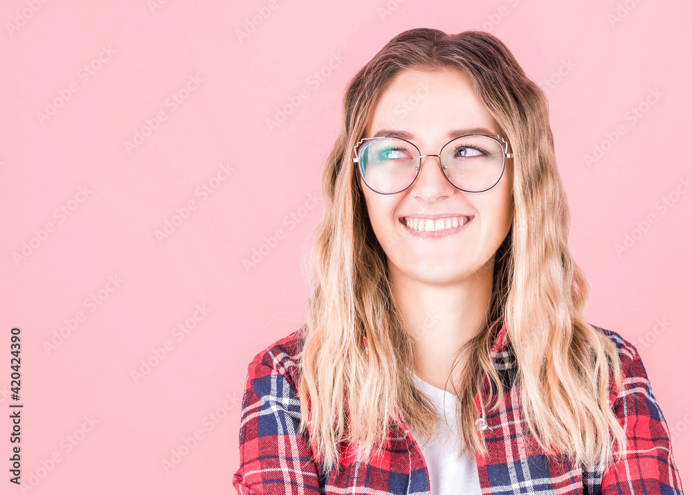 cheerful happy young beautiful curly girl, in glasses and a checkered red shirt looking to the side, folded her arms, smiling, laughing on a pink background.