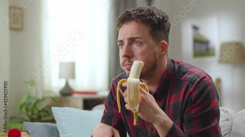Young handsome man eats banana and watching tv at home in the living room.