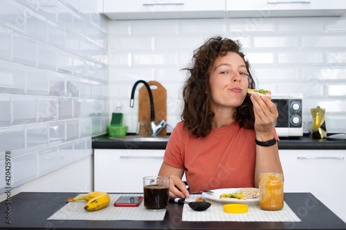Young brunette woman enjoying breakfast in white kitchen at home,sitting at table chewing healthy bread with avocado and peanut butter looking at camera with joy.Morning routine.Vegetarianism concept