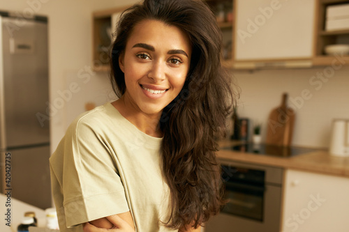 People, relaxation, rest and leisure concept. Close up shot of adorable young Hispanic female in casual comfy t-shirt posing in kitchen interior, spending day at home, looking at camera with smile