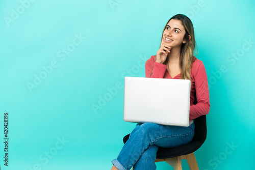 Young caucasian woman sitting on a chair with her pc isolated on blue background thinking an idea while looking up © luismolinero